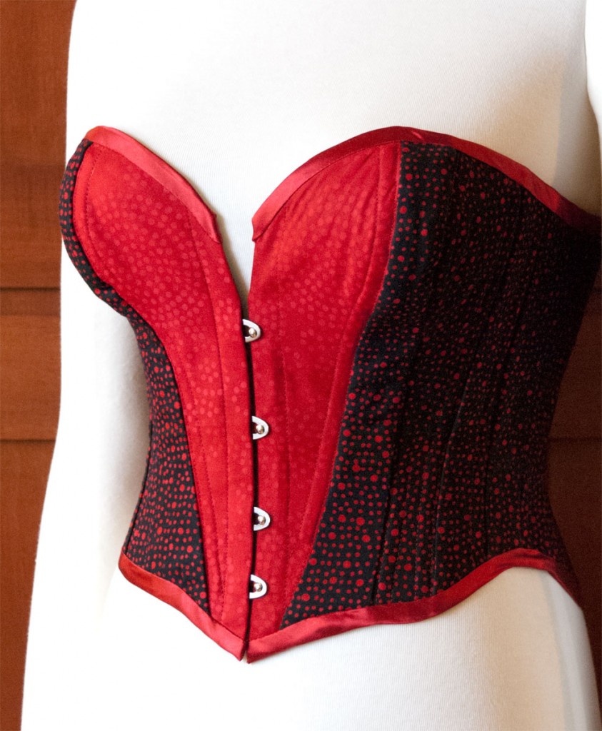 An overbust corset made with red and black cotton batik fabric for outside and lining, trimmed with silk satin. It has a steel busk in front, spiral steel bones and two layers of cotton coutil on the inside.